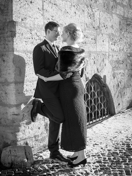 Hochzeitsportraet_Erfurt_Mauer_Verliebtes-Brautpaar_Michael-Schalansky Schwarz-Weiss-Porträt eines verliebten Brautpaares, das sich eng umschlungen an eine raue Steinmauer lehnt. Der Mann im dunklen Anzug, die Frau in einem dunklen, eleganten Kleid mit Pelzstola. Sie schauen sich lächelnd in die Augen. Der Boden ist mit Kopfsteinpflaster und etwas Schnee oder Frost bedeckt. Das Bild vermittelt Romantik und Nähe und ist ein Beispiel für Hochzeitsfotografie von Michael Schalansky in Erfurt.